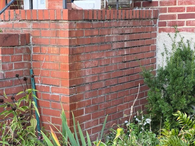 Brick wall with horizontal cracks, part of a building's entrance. Green foliage surrounds the base.