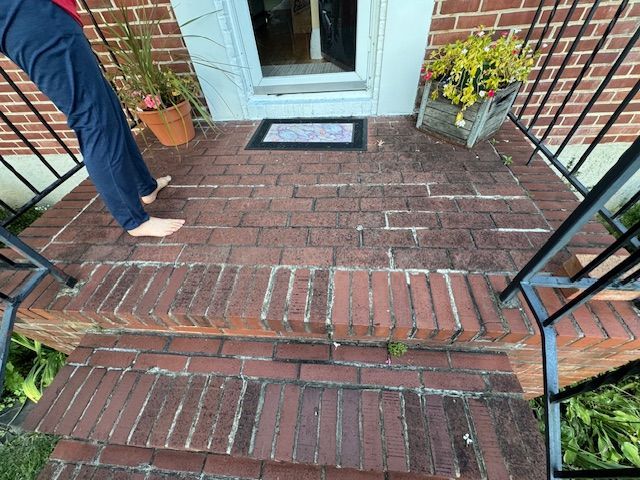 Brick porch with steps; person's legs visible. Pots of flowers, a welcome mat, and a doorway are present.