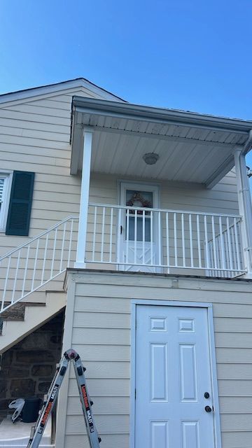 Beige house with porch and white railing, white door, and green shutters. A ladder leans against the building.