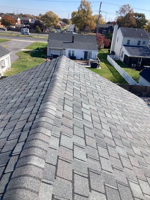 Gray shingle roof, angled view. Houses, green lawns, and blue sky visible in the background.