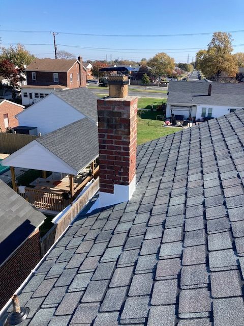 Brick chimney on a gray shingle roof, surrounded by houses under a blue sky.
