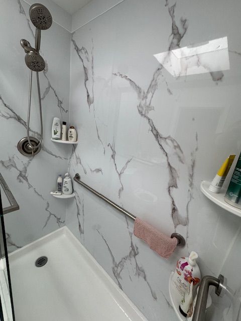 Shower with white marble-patterned walls, grab bar, showerhead, and toiletries on shelves.