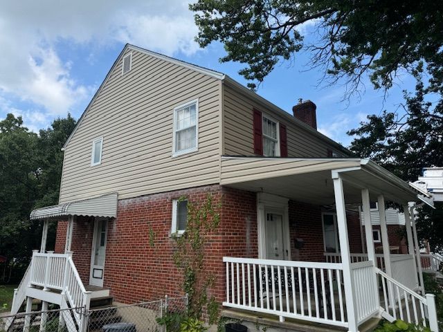 Two-story house with brick base, tan siding, white porch, and red shutters under a cloudy blue sky.