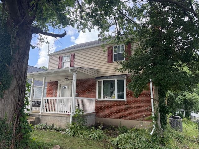 Two-story house with a porch. Brick and tan siding, red shutters, and white trim. Green trees and sky visible.