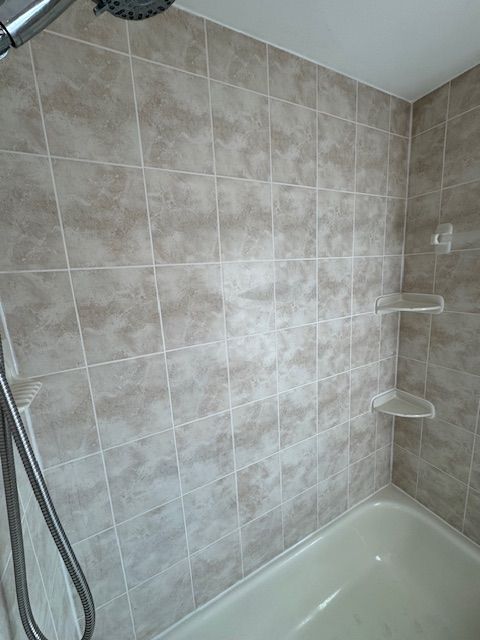 Shower with beige-toned tile walls and a white bathtub. Two corner shelves are visible.