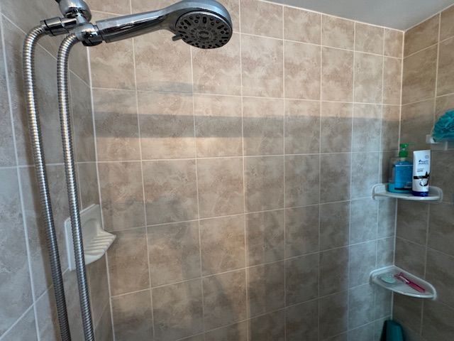 Shower stall with beige tile, showerhead, and a shelf with toiletries.