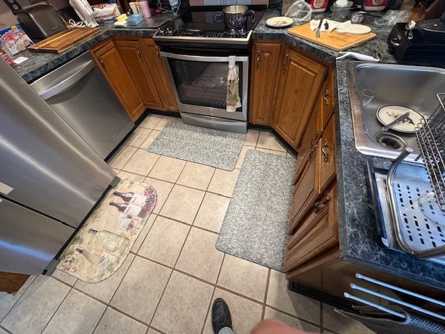 Kitchen with stainless steel appliances, wooden cabinets, and tile floor with small rugs.