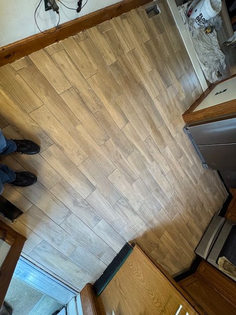 Light wood-look flooring installed in a kitchen. A person's feet, appliances, and cabinetry are visible.