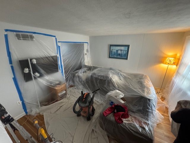 Room being prepped for painting: furniture and electronics covered in plastic sheeting. Blue tape outlines doorways.