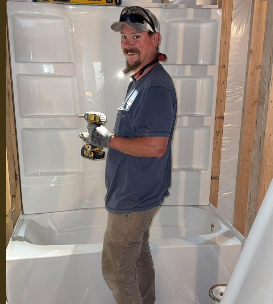 Man installing a white bathtub and shower surround with a drill in a construction setting.