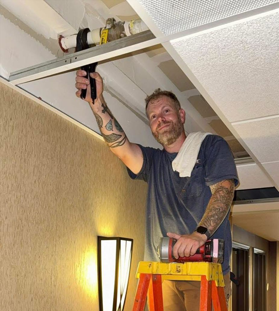 Man on step stool working on ceiling tiles, holding pliers and a flashlight. Hallway setting.