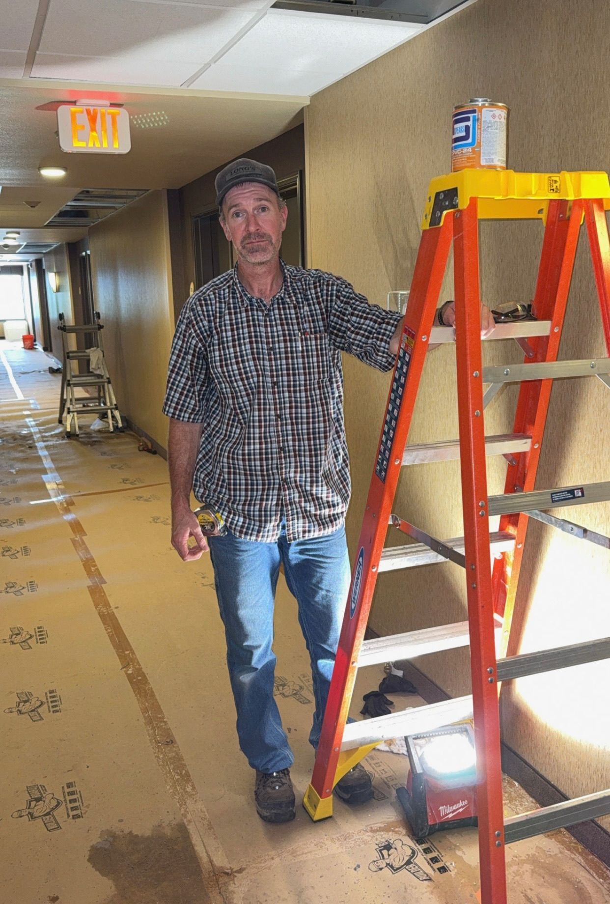 Man in plaid shirt and jeans stands by ladder in hallway under construction; paint can on top.