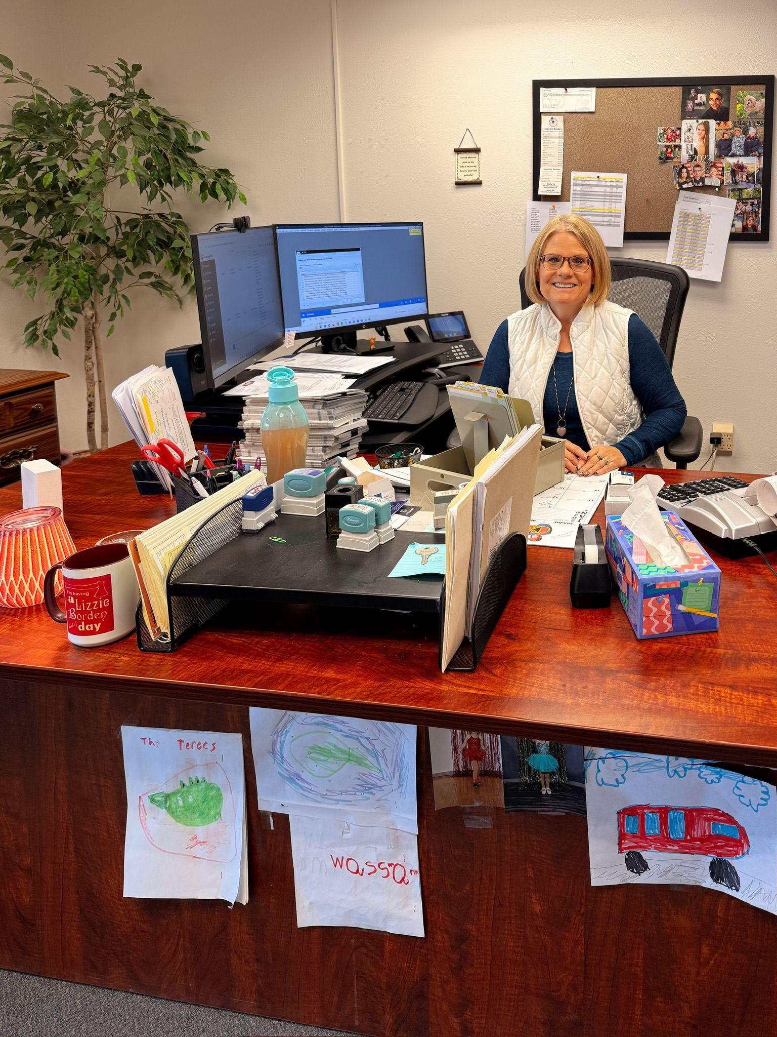 Woman seated at a messy desk with two monitors. Drawings are taped to the desk front.