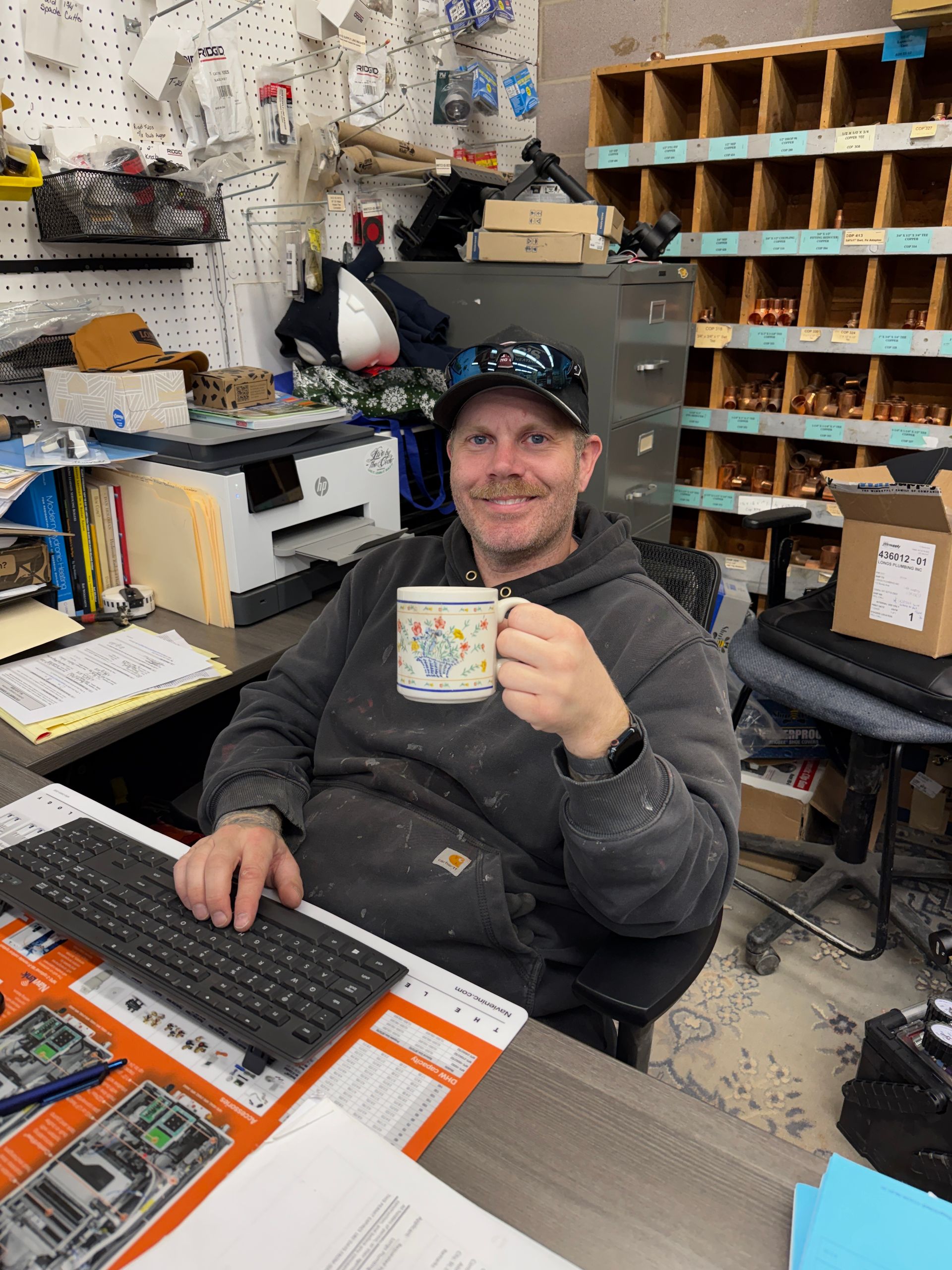 Man in workshop holding mug, smiling, seated at desk with keyboard. Workshop setting with shelves.
