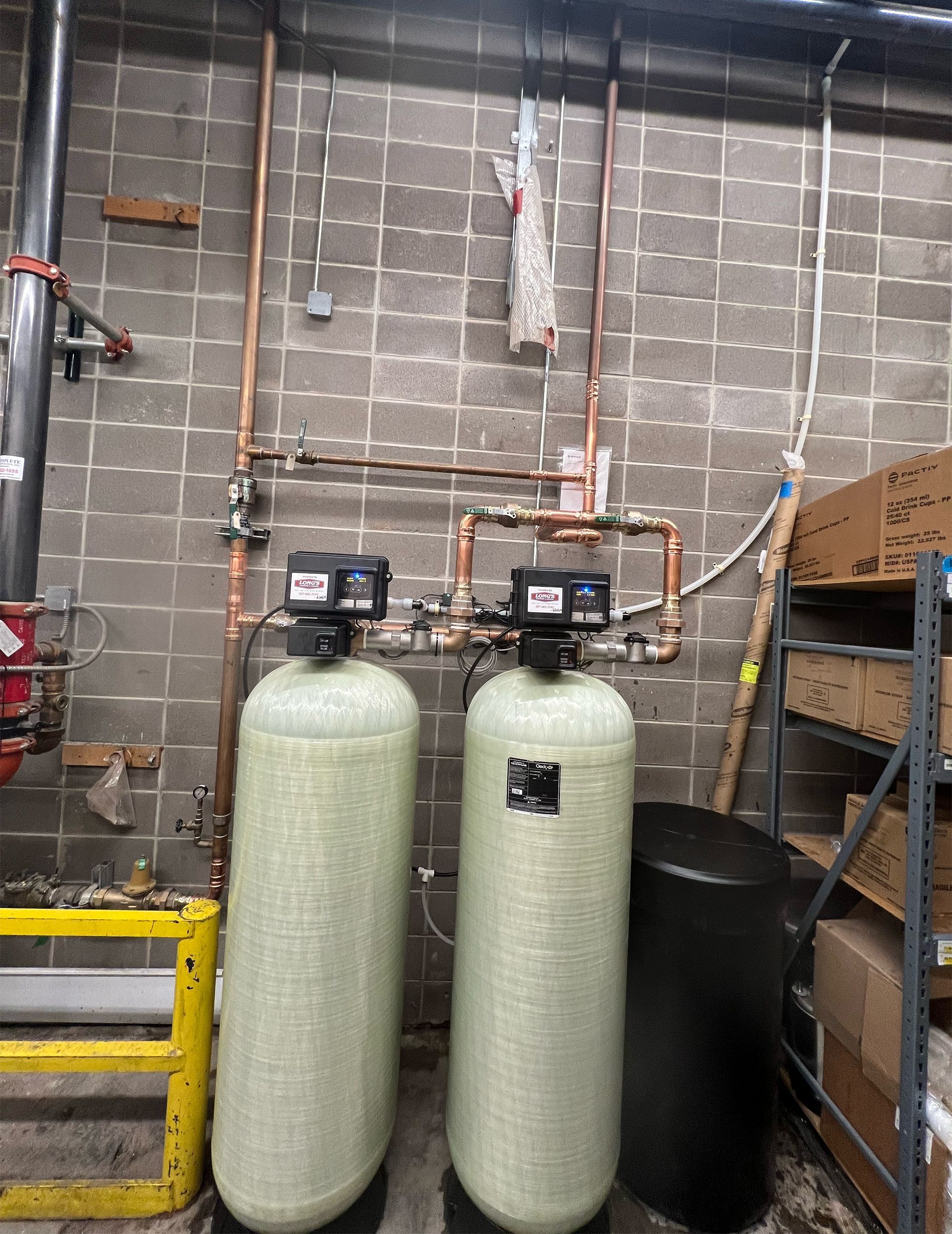 Two tall industrial water softening tanks with electronic control heads mounted against a concrete block wall.