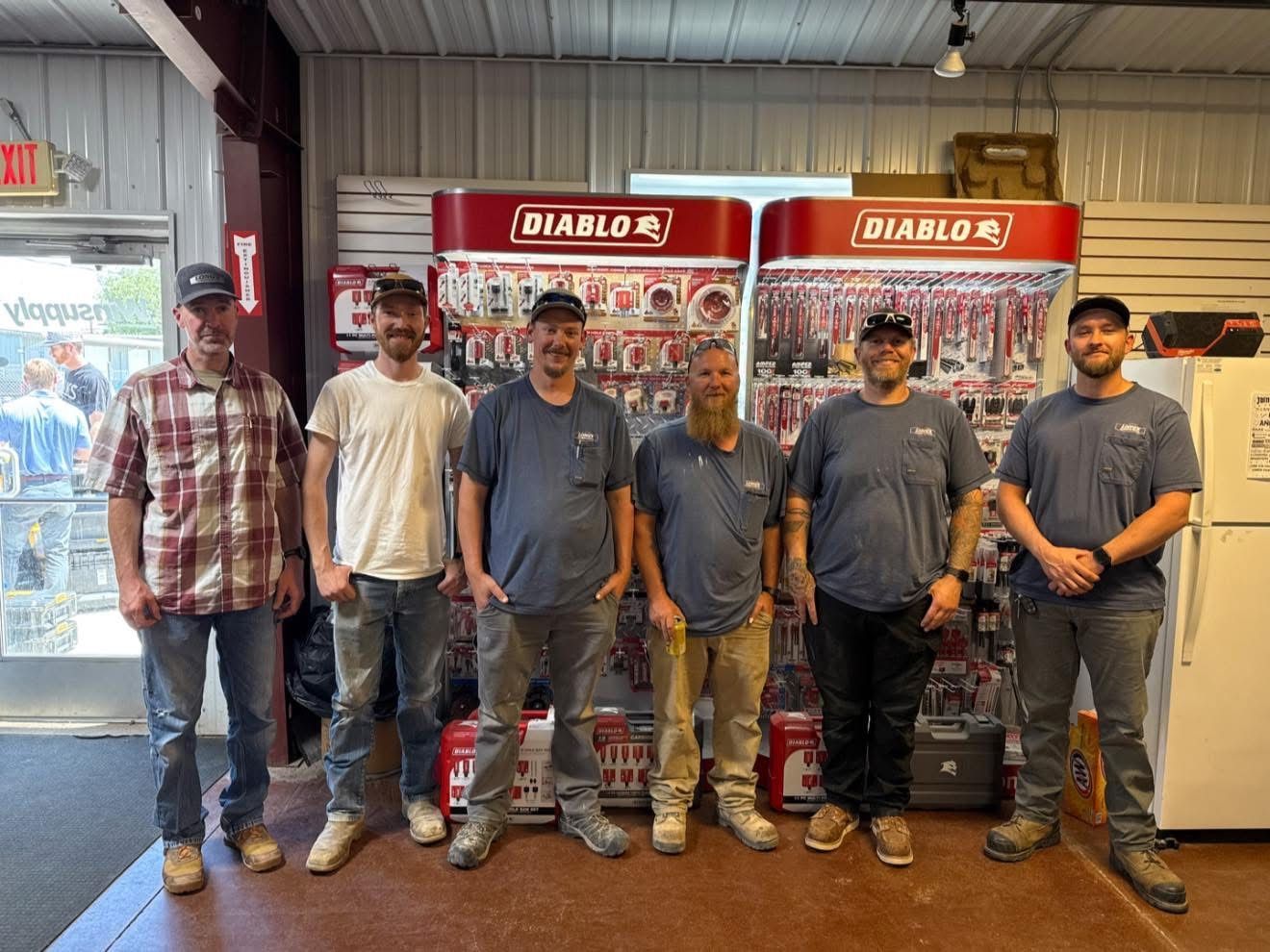 Six men pose inside a hardware store, in front of tool displays.