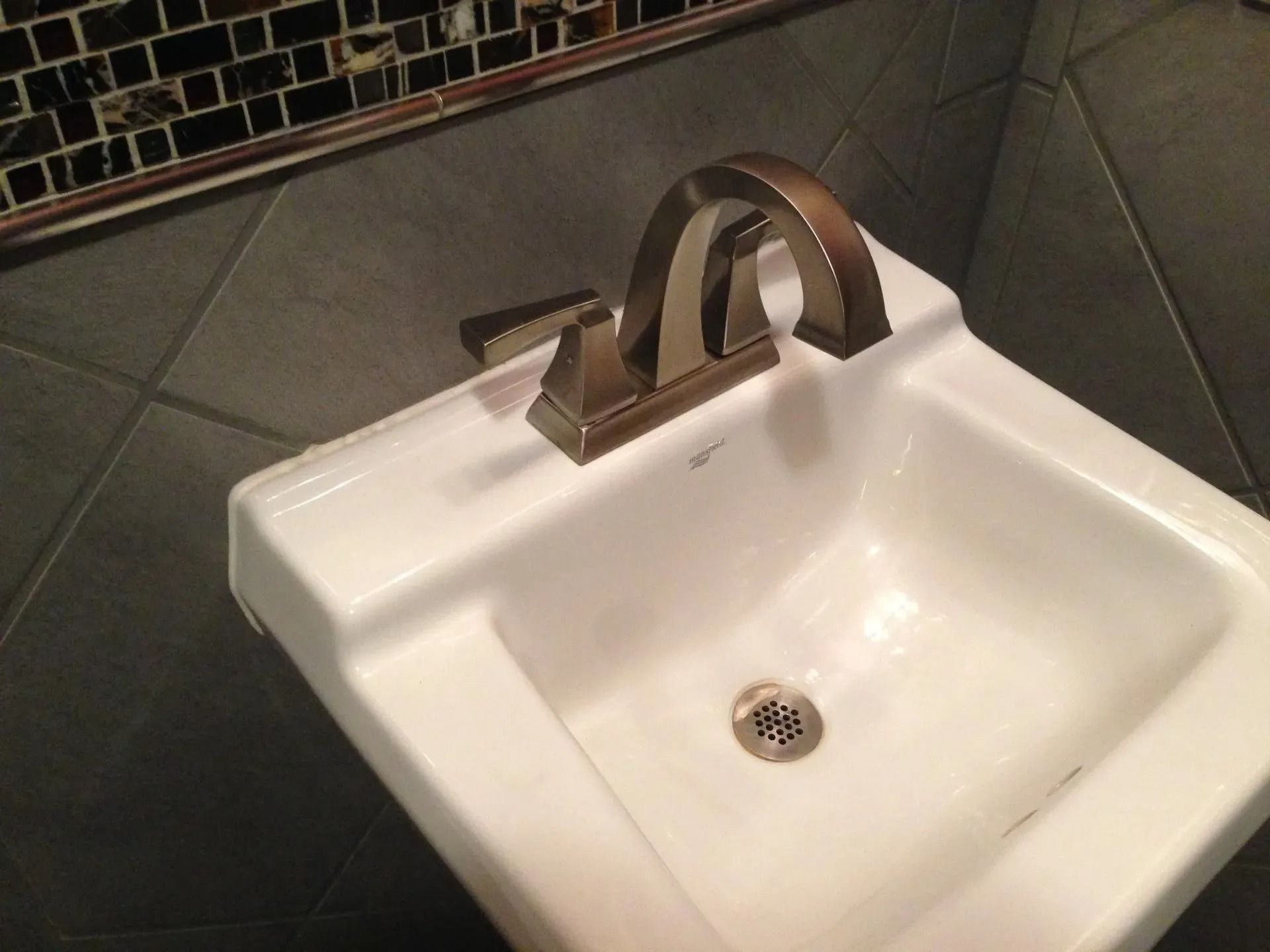 White bathroom sink with a bronze faucet against a gray tiled wall.