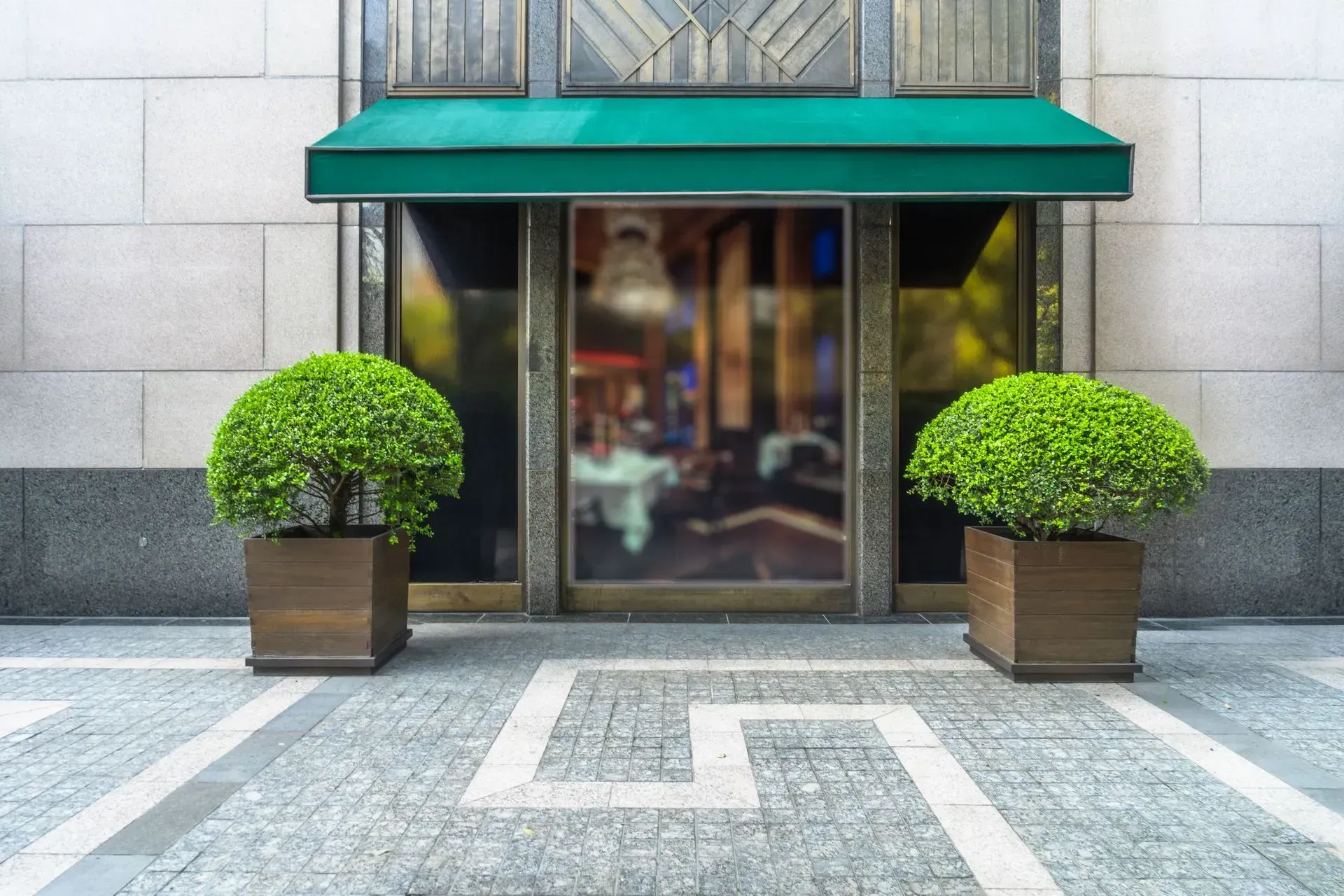 Storefront entrance with teal awning, glass doors, and two trimmed potted shrubs