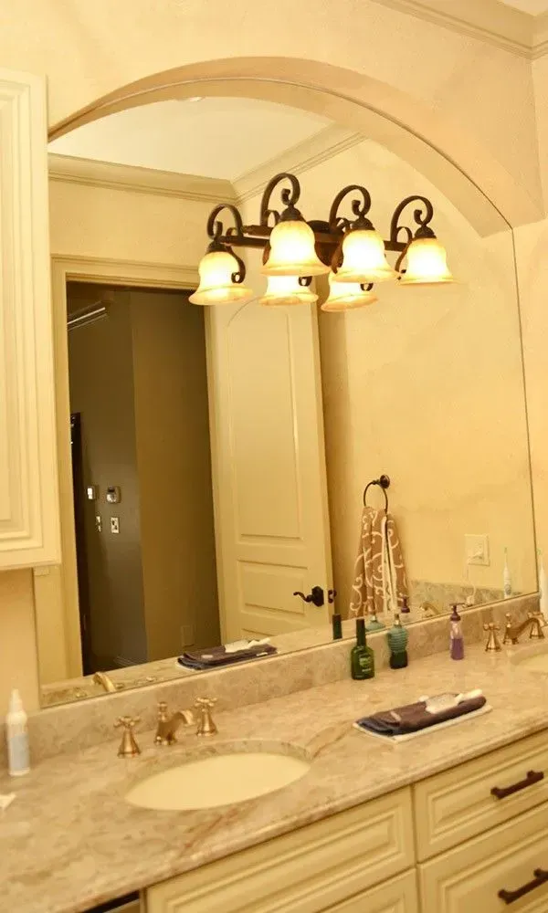 Warm bathroom vanity with granite countertop, dual sinks, large mirror, and lit chandelier in cream tones