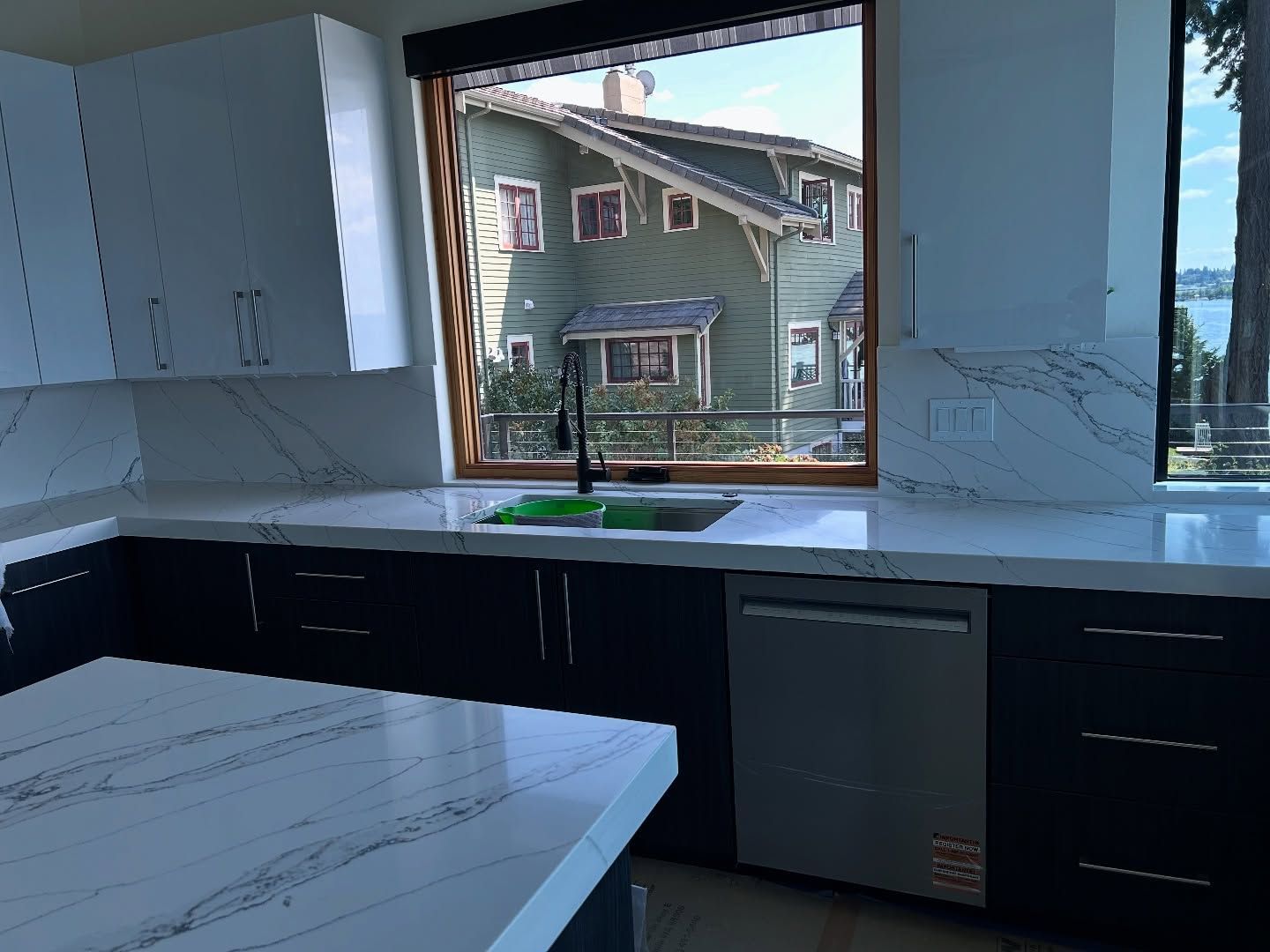 Modern kitchen with white countertops and dark cabinets, overlooking a house and water.
