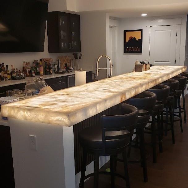 Lit-up countertop bar with black stools in a home basement. Dark wood cabinets and a sink are in the background.