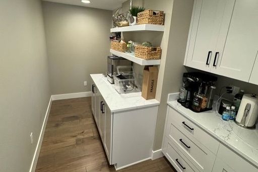 Coffee station in a kitchen, with white cabinets, countertop, shelves and appliances. Wooden floor and tan walls.