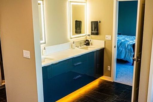 Modern bathroom with a navy blue vanity, lit mirrors, and a bedroom visible in the doorway.