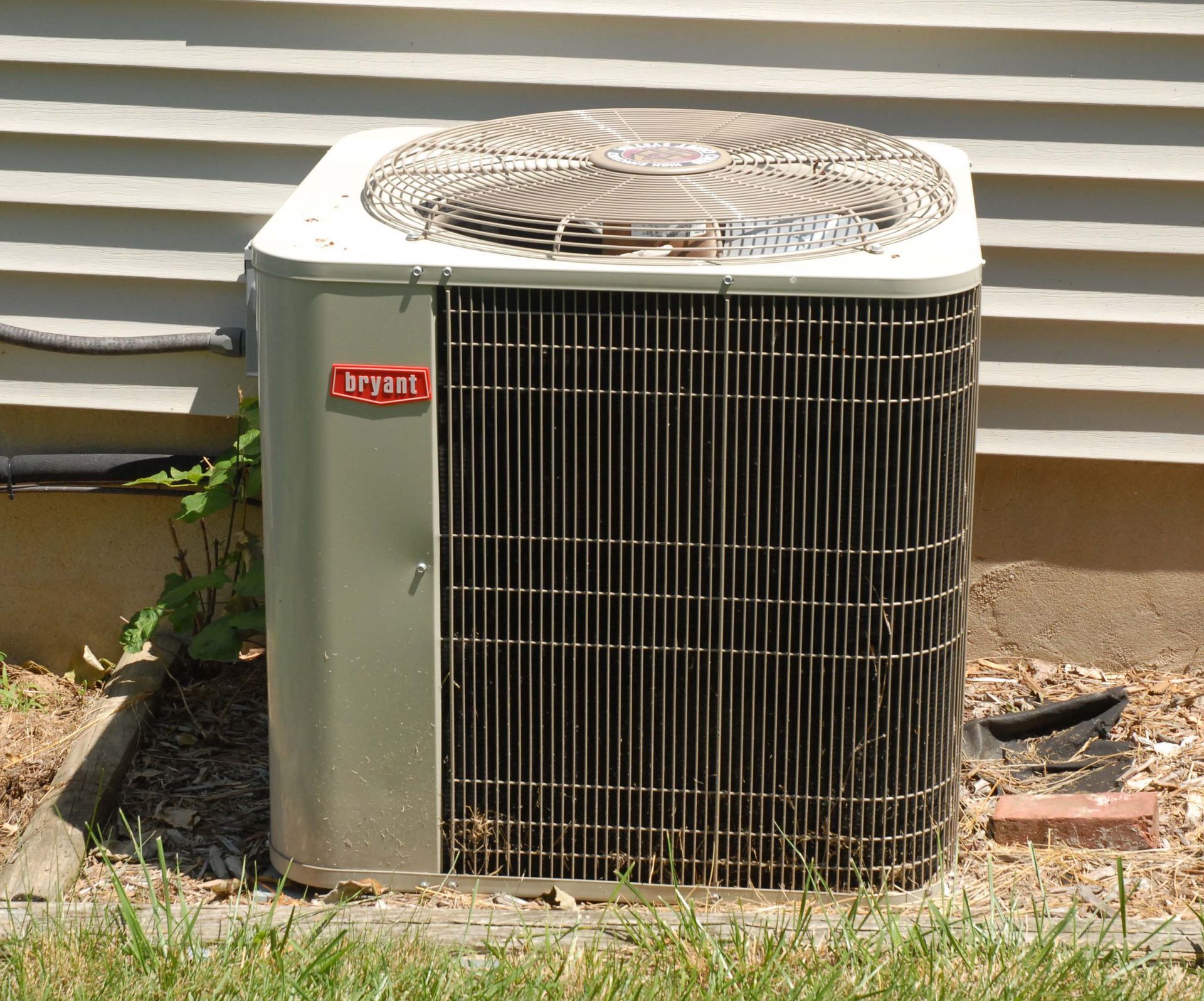 Old outdoor HVAC unit beside a house wall, with a fan on top and grassy ground below