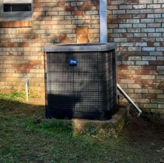Outdoor HVAC unit beside a brick wall with a metal conduit and patchy grass below.