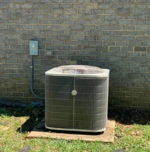 Outdoor air conditioning unit beside a brick wall on a concrete pad, with grass in front.