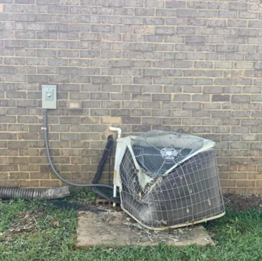 Damaged outdoor air conditioner unit against a brick wall, with a bent metal cage and cable attached.