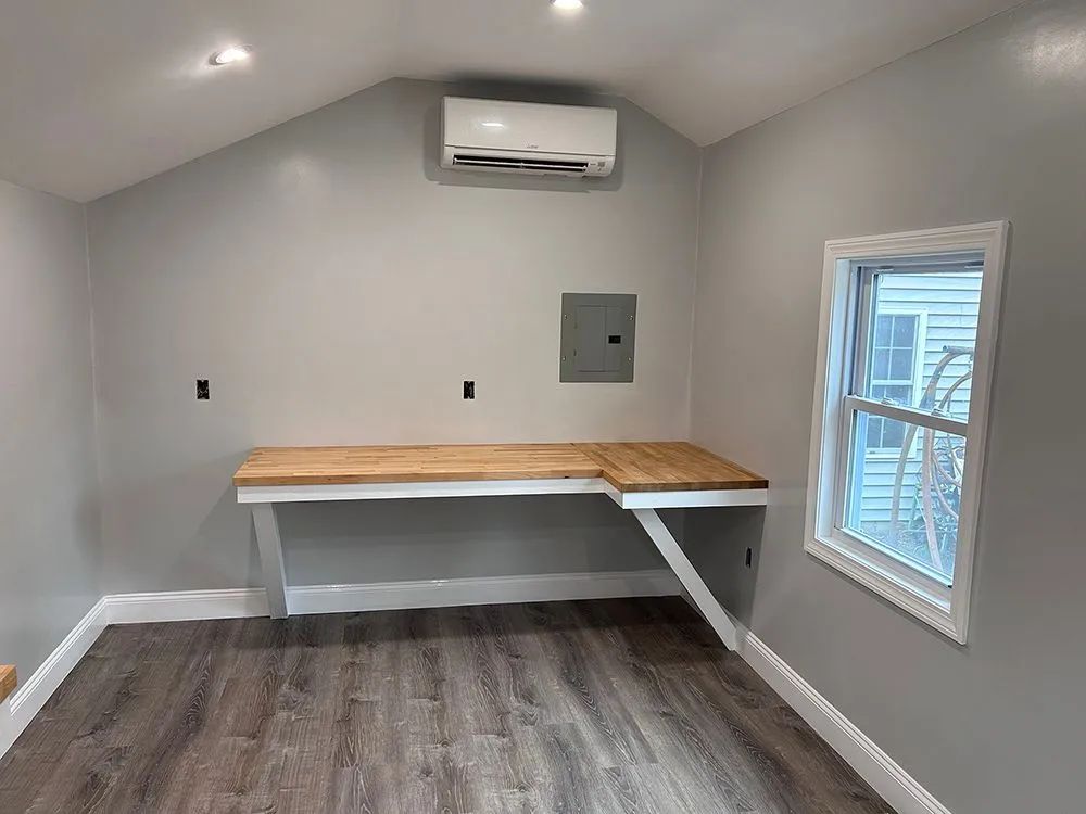 A small, light-gray room with a built-in wooden desk, window, and air conditioner.