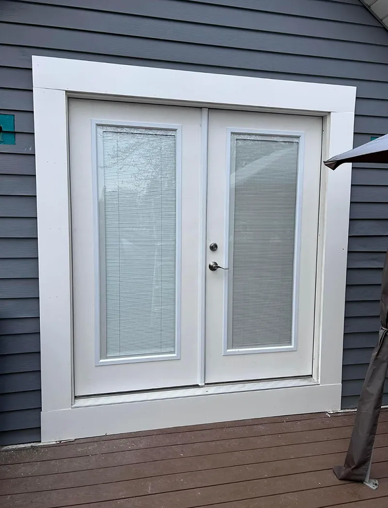 White French doors with blinds, trimmed in white, set into a grey-sided building. Brown deck visible.