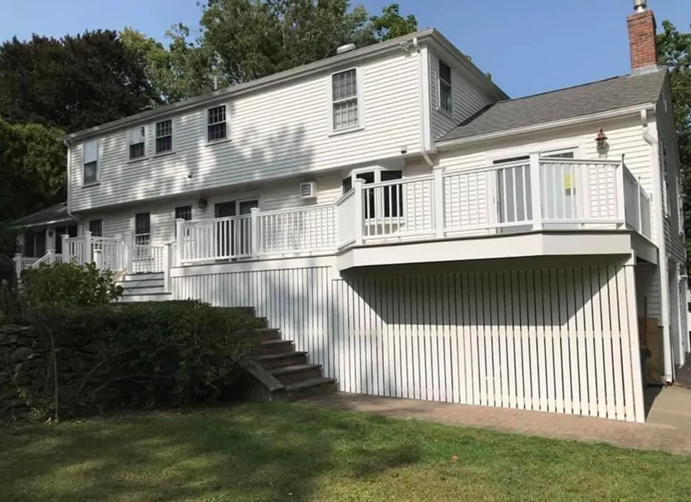 Back view of a white, multi-level house with a large deck and stairs, set on a grassy lawn.