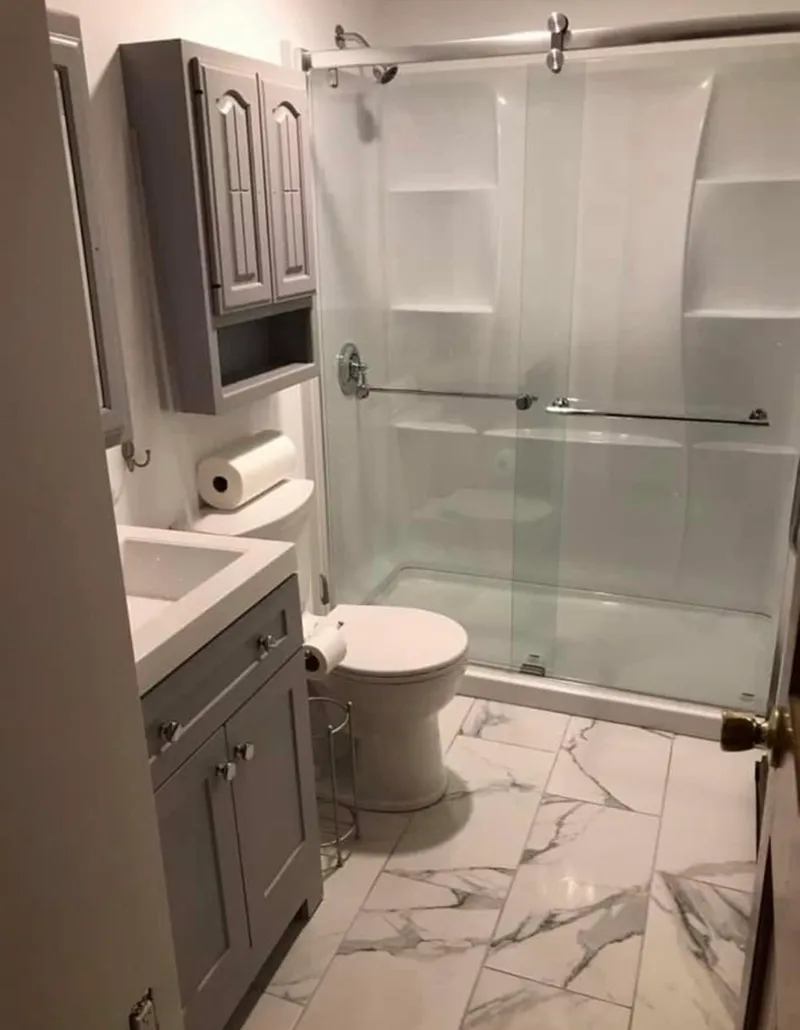 Bathroom with gray vanity, toilet, and shower. White marble-look floor tiles. Gray upper cabinet.