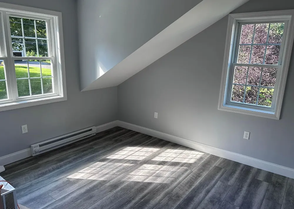 Gray-walled room with two windows and wood-look flooring. Light streams across the floor from the windows.