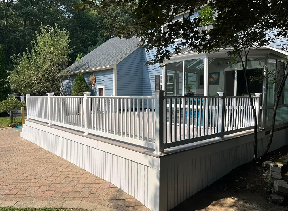 White deck with railing, connected to a blue house with sunroom. Overlooks a brick patio.