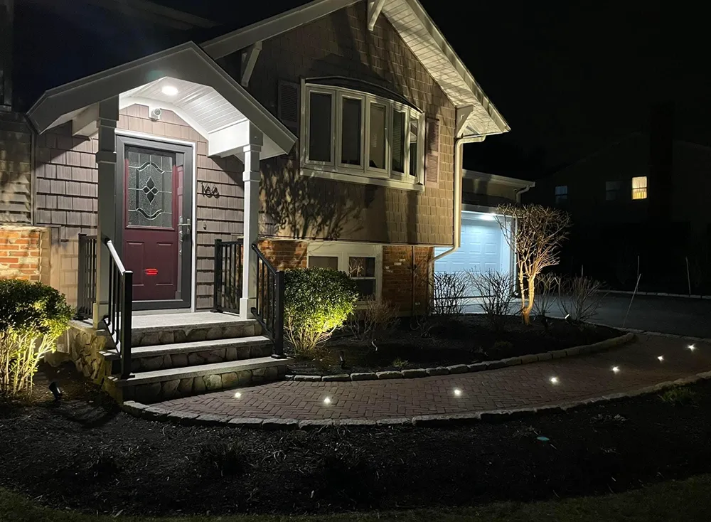 House exterior at night with illuminated walkway, porch, and landscaping. Dark brown siding, red door, glowing lights.