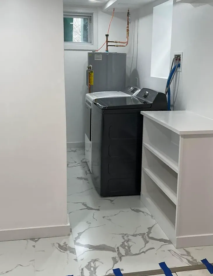 Laundry room with washing machine, dryer, water heater, and shelving. White walls, marble-look floor.