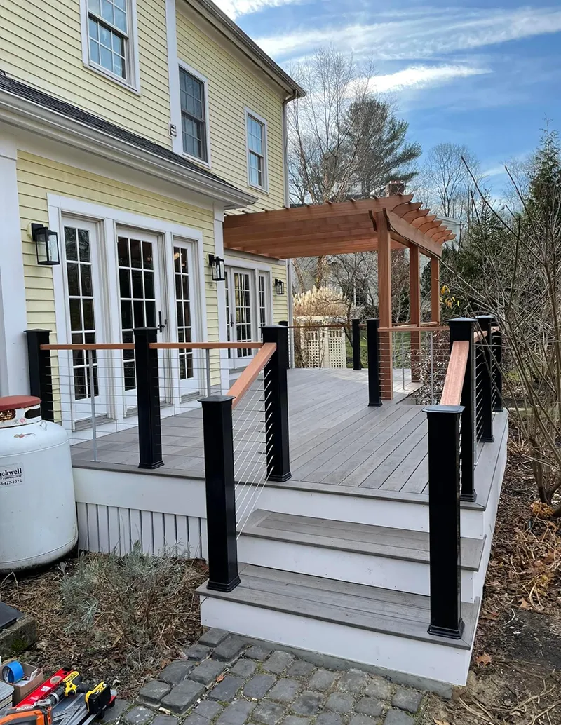 Wooden deck with a pergola, black posts, and tan railing next to a yellow house with a lake visible in the background.