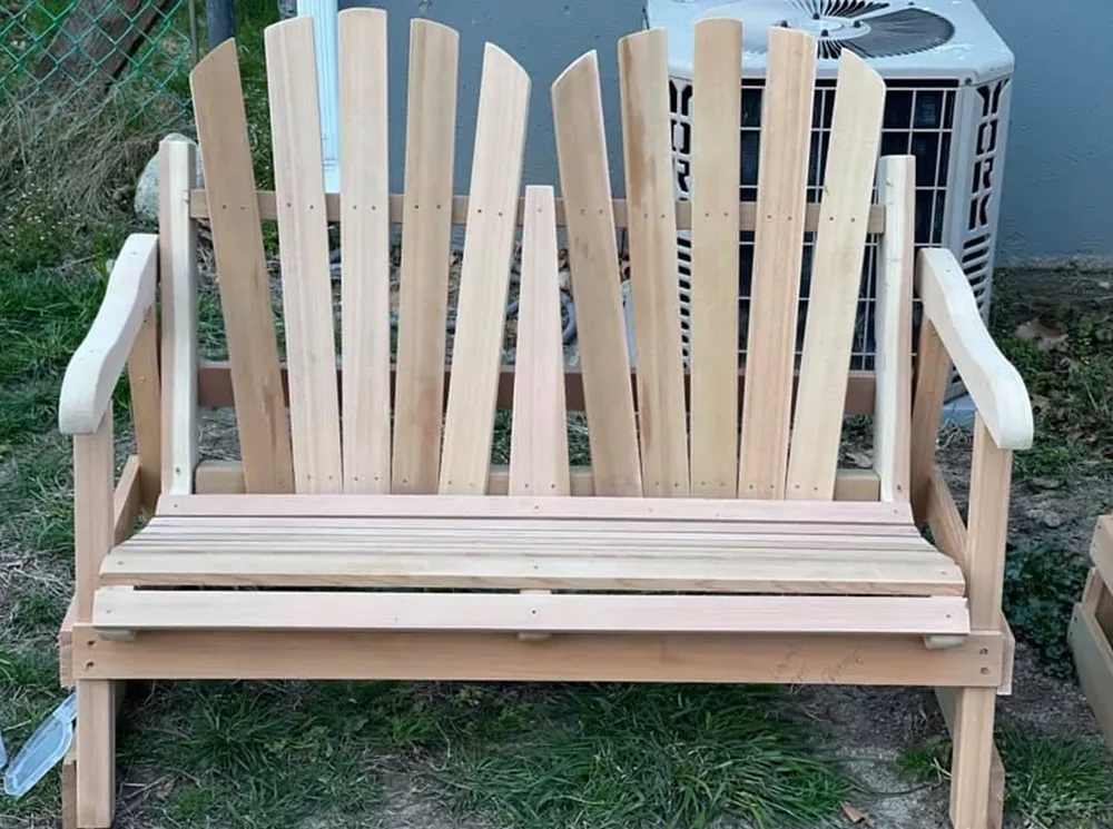 Wooden outdoor double bench. Light brown wood, slat backrests, armrests. Outside, grass background.