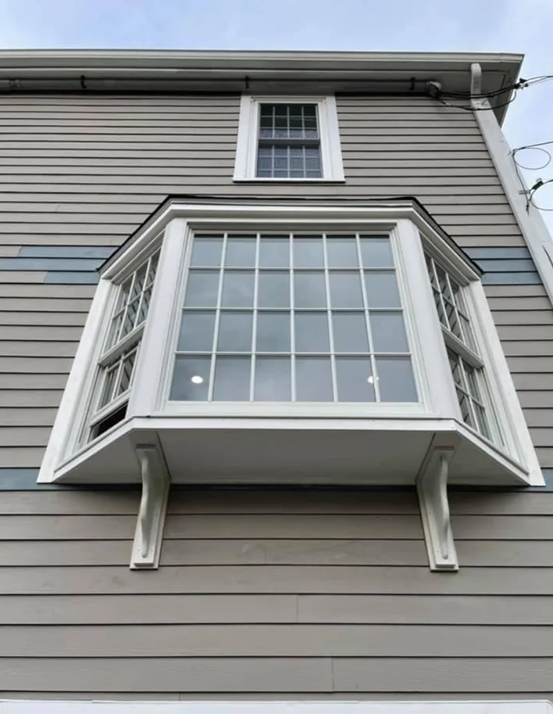 Bay window with white trim on a light gray house; small window above.