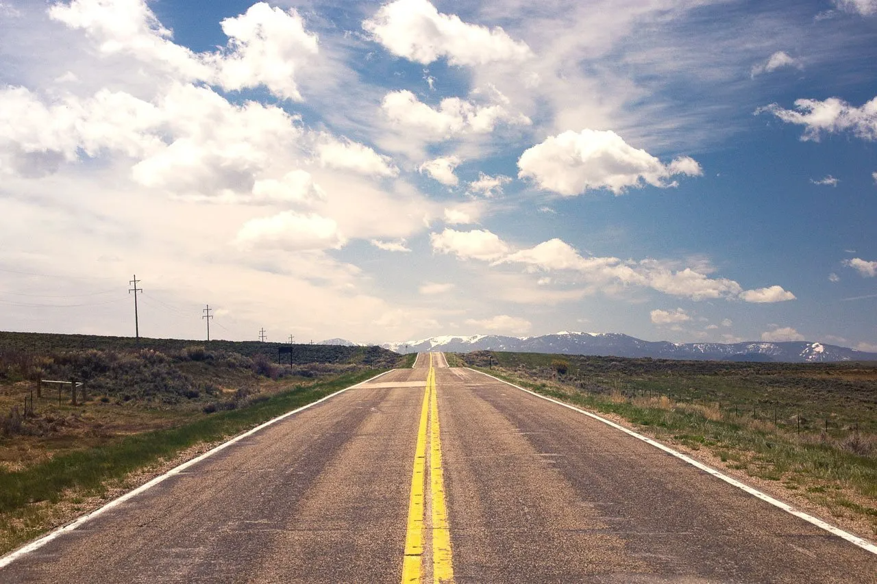 Open road with yellow lines under a blue sky dotted with puffy clouds, framed by fields and distant mountains.