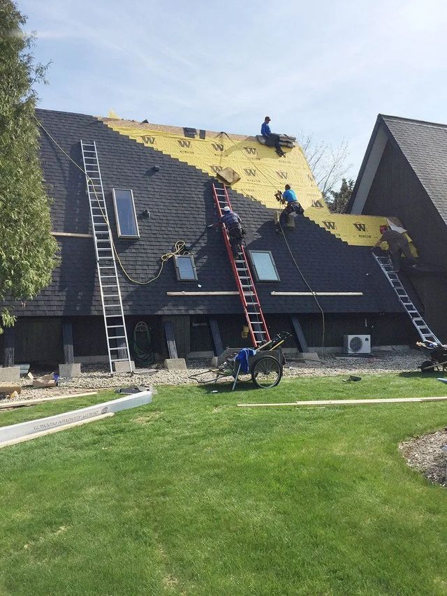 Roofers working on a house roof, removing old shingles and installing new ones; ladders, bright sky.