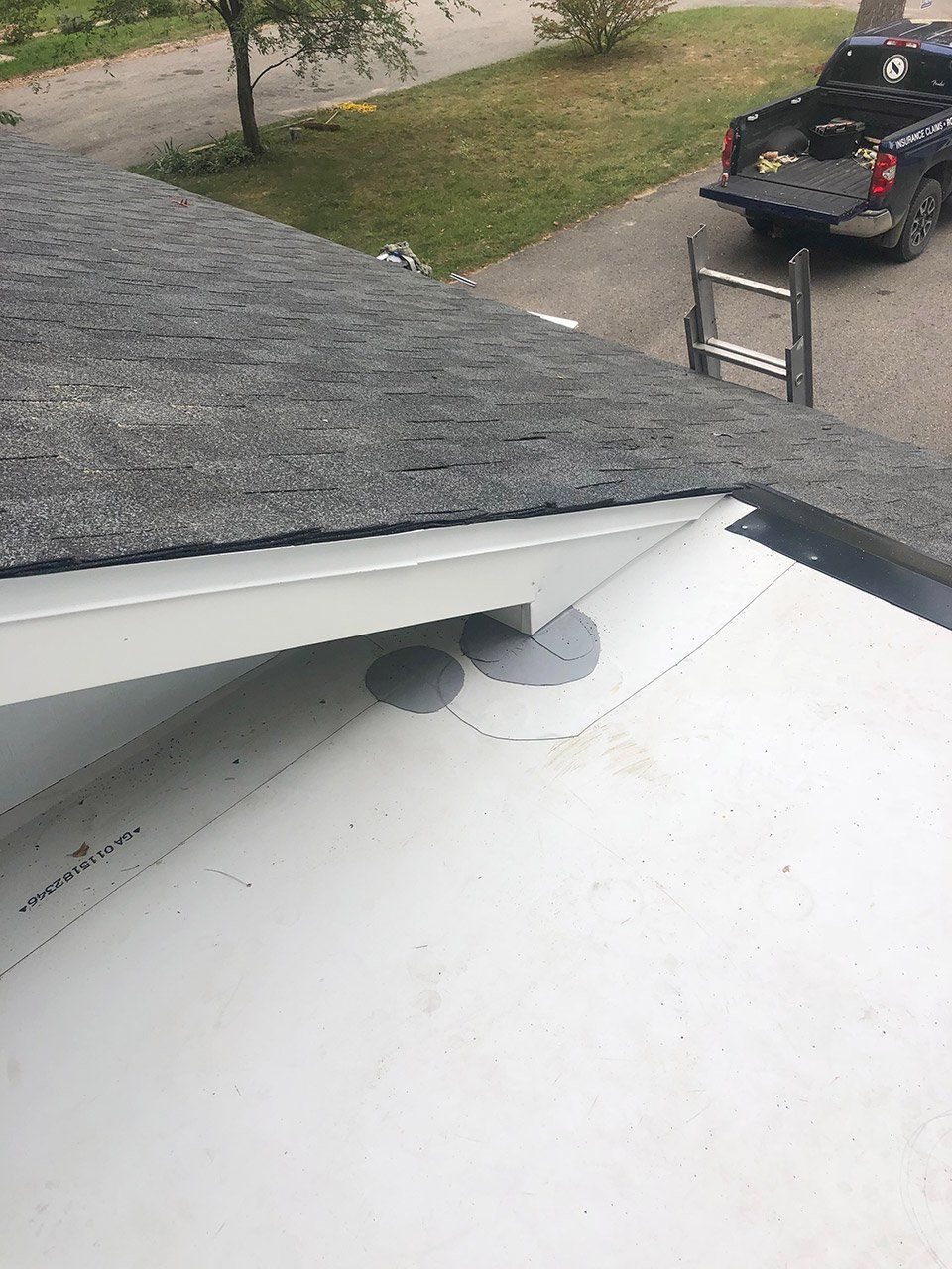 Overhead view of a roof corner with black shingles, white trim, and a gray-covered vent, with a street and truck in the background.
