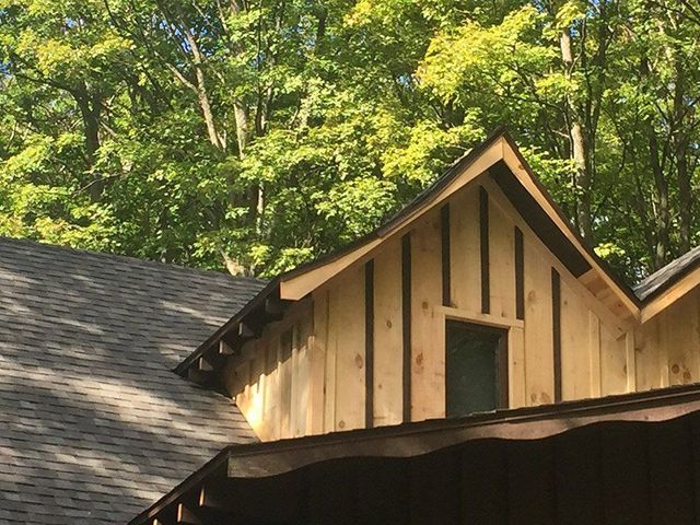 Gabled wooden dormer on a roof with a dark shingled finish. Trees are in the background.