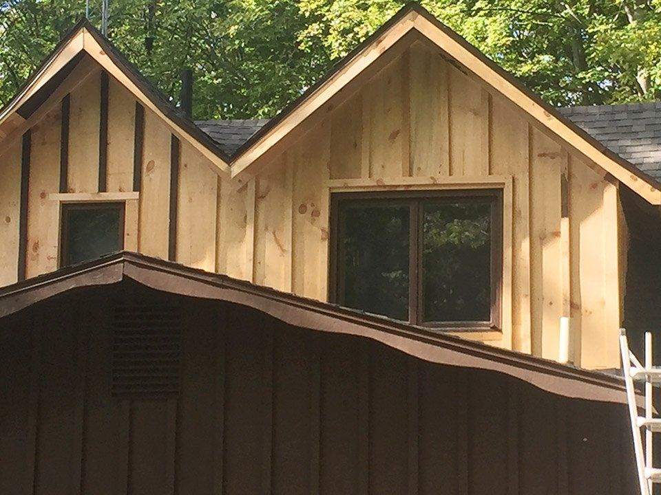 Brown and natural wood house exterior with two gabled dormers and dark windows, set against a green tree backdrop.