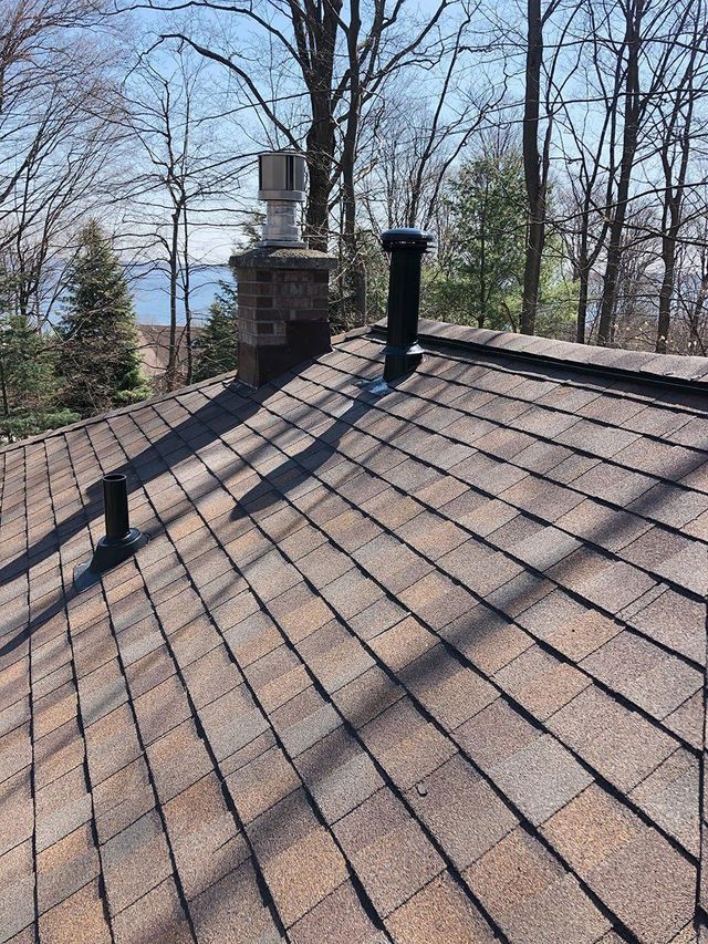 Brown shingled roof with chimneys and vents against a backdrop of trees and a body of water.