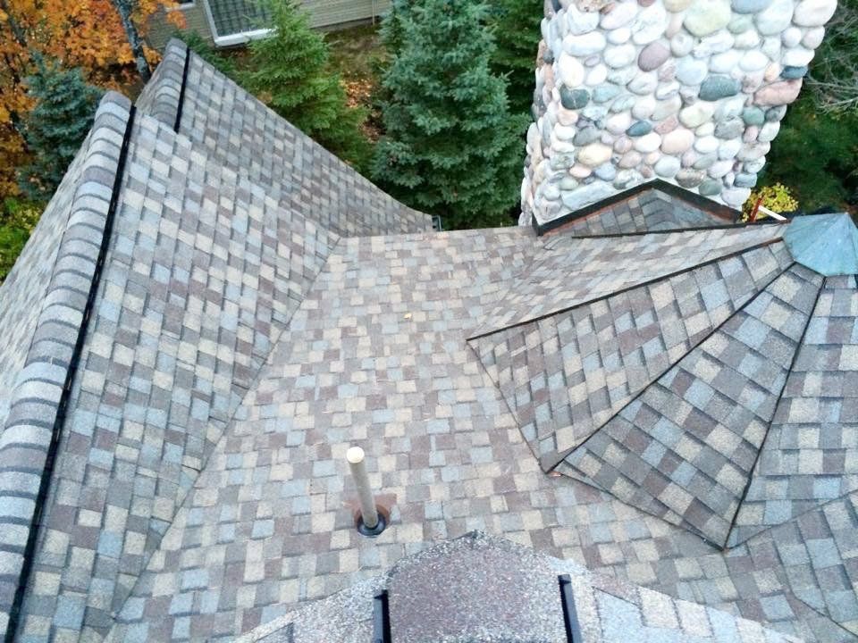Overhead view of a house roof with multicolored shingles, a stone chimney, and trees in the background.