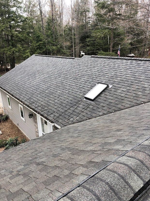 Overhead view of two gray shingled roofs, one with a skylight, surrounded by trees.